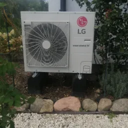 Zewnętrzna jednostka pompy ciepła LG Therma V, zamontowana na betonowych podstawach w ogrodzie, z widocznymi kamieniami i roślinnością w tle.
