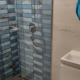 Nowoczesna kabina prysznicowa z niebieskimi, prostokątnymi płytkami imitującymi cegłę, szklana ściana, chromowana armatura i odpływ liniowy w podłodze. Obok umywalka z szafką w kolorze białym.