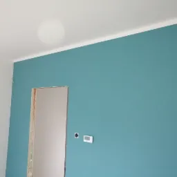 Świeżo pomalowane wnętrze z turkusową ścianą i białym sufitem, widoczny otwór drzwiowy w stanie surowym i regulator temperatury na ścianie.