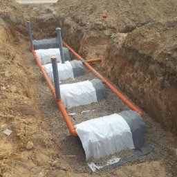 Drenaż rozsączający w wykopie, rury pomarańczowe i szare, geowłóknina biała i szara, obsypane żwirem, widoczne ściany wykopu z ziemi.