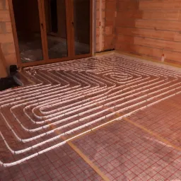 Montaż ogrzewania podłogowego w Drezdenku: białe rury ułożone w zygzak na czerwonej siatce, widoczne surowe ściany z cegły i okno.