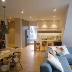 Jasne, przestronne wnętrze domu z drewnianą podłogą, nowoczesną kuchnią z barkiem i białymi szafkami, jadalnią z drewnianym stołem i szarą sofą z poduszkami.