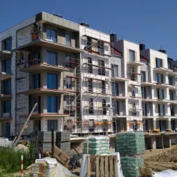 Węgorzewo: Budynek wielorodzinny w trakcie prac elewacyjnych, widoczne rusztowania, warstwa izolacyjna na części fasady oraz składowane materiały budowlane.