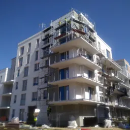 Wielopiętrowy budynek mieszkalny w trakcie prac elewacyjnych, widoczne rusztowania, pracownicy w kamizelkach odblaskowych, częściowo ocieplona ściana i materiały budowlane na ziemi.