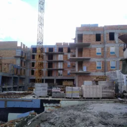 Budowa wielopiętrowego budynku z cegły, widoczny żuraw budowlany i betoniarka, materiały budowlane na paletach