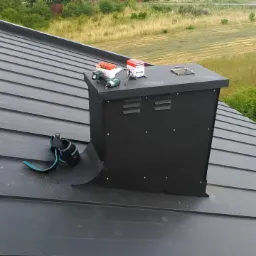 Czarny komin wentylacyjny na dachu z blachy trapezowej, narzędzia pracy i puszki z uszczelniaczem leżące na górnej krawędzi komina, widok z góry na tle łąk.