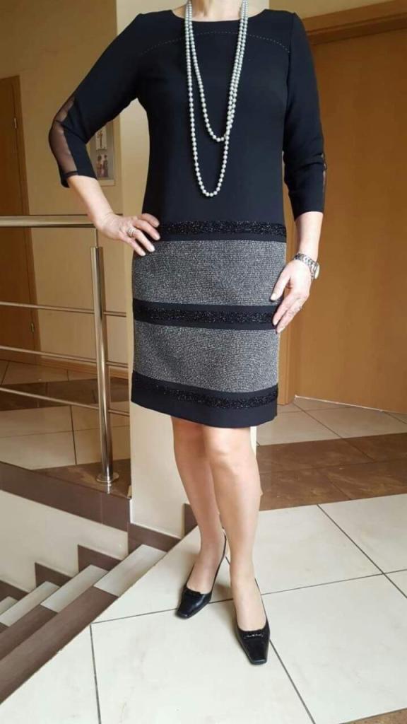 Kobieta prezentuje czarną sukienkę z szarym, poziomym wzorem, uzupełnioną o długi naszyjnik z pereł i czarne lakierowane buty na niskim obcasie. Sukienka posiada rękawy 3/4 z siateczkowymi wstawkami.