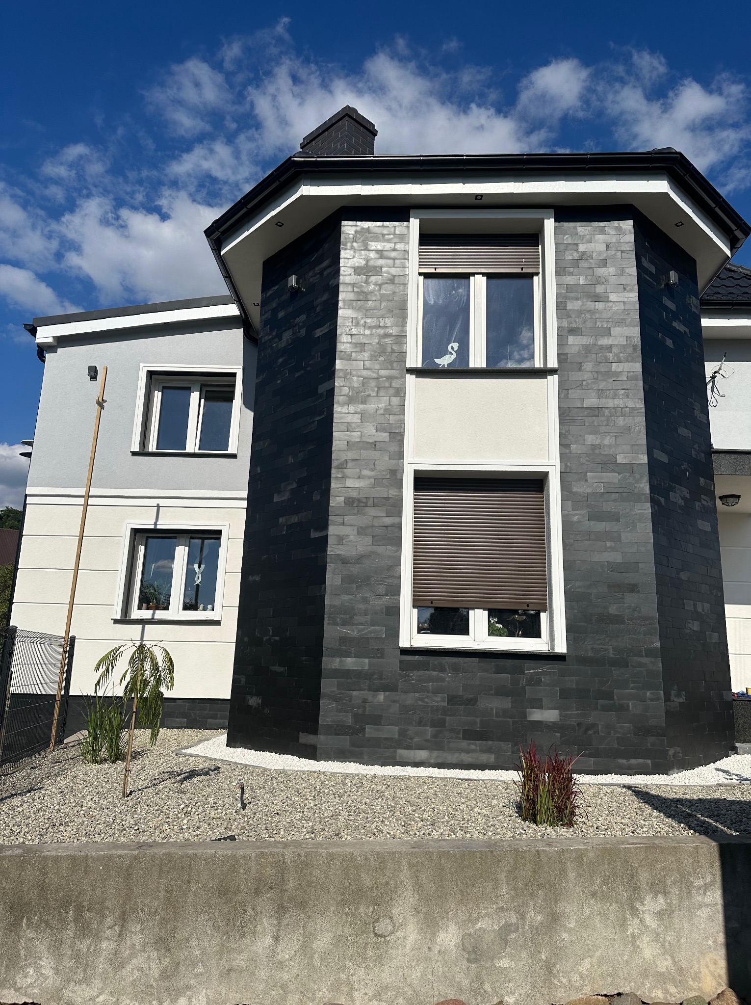 Dwukondygnacyjny dom z elewacją w odcieniach szarości i czerni, wyróżniający się cylindryczną częścią pokrytą czarną cegłą, z oknami z roletami i dekoracją okienną w kształcie łabędzia, otoczony...