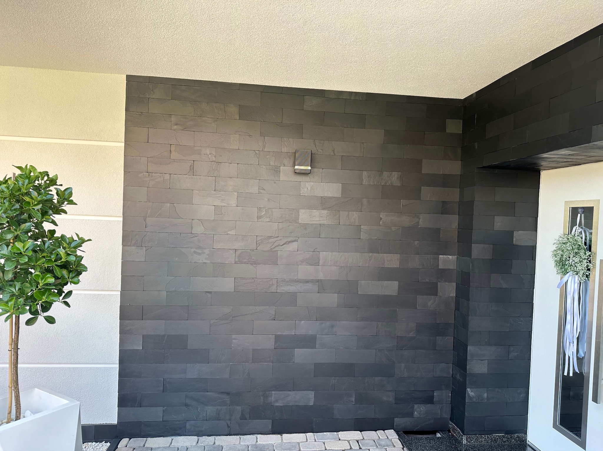 Nowoczesna elewacja wykończona czarnym kamieniem łupkowym w kształcie cegiełek, z widoczną lampą ścienną, obok drzwi z dekoracją kwiatową i donica z drzewkiem.