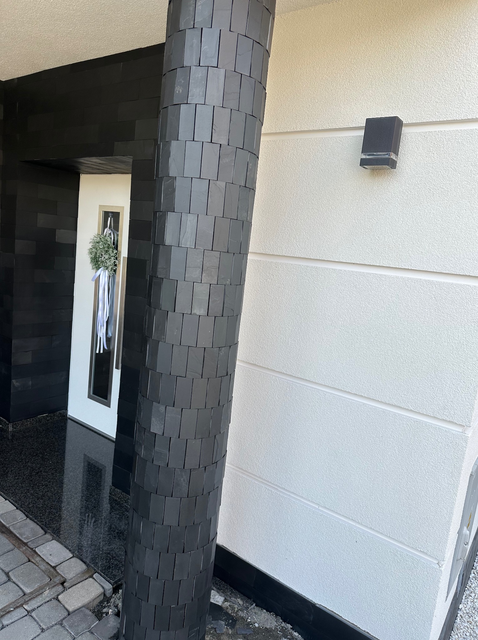 Nowoczesna elewacja domu z czarną okładziną z płytek na filarze i fragmencie ściany, biała strukturalna ściana z lampą zewnętrzną, wieniec nad drzwiami wejściowymi.