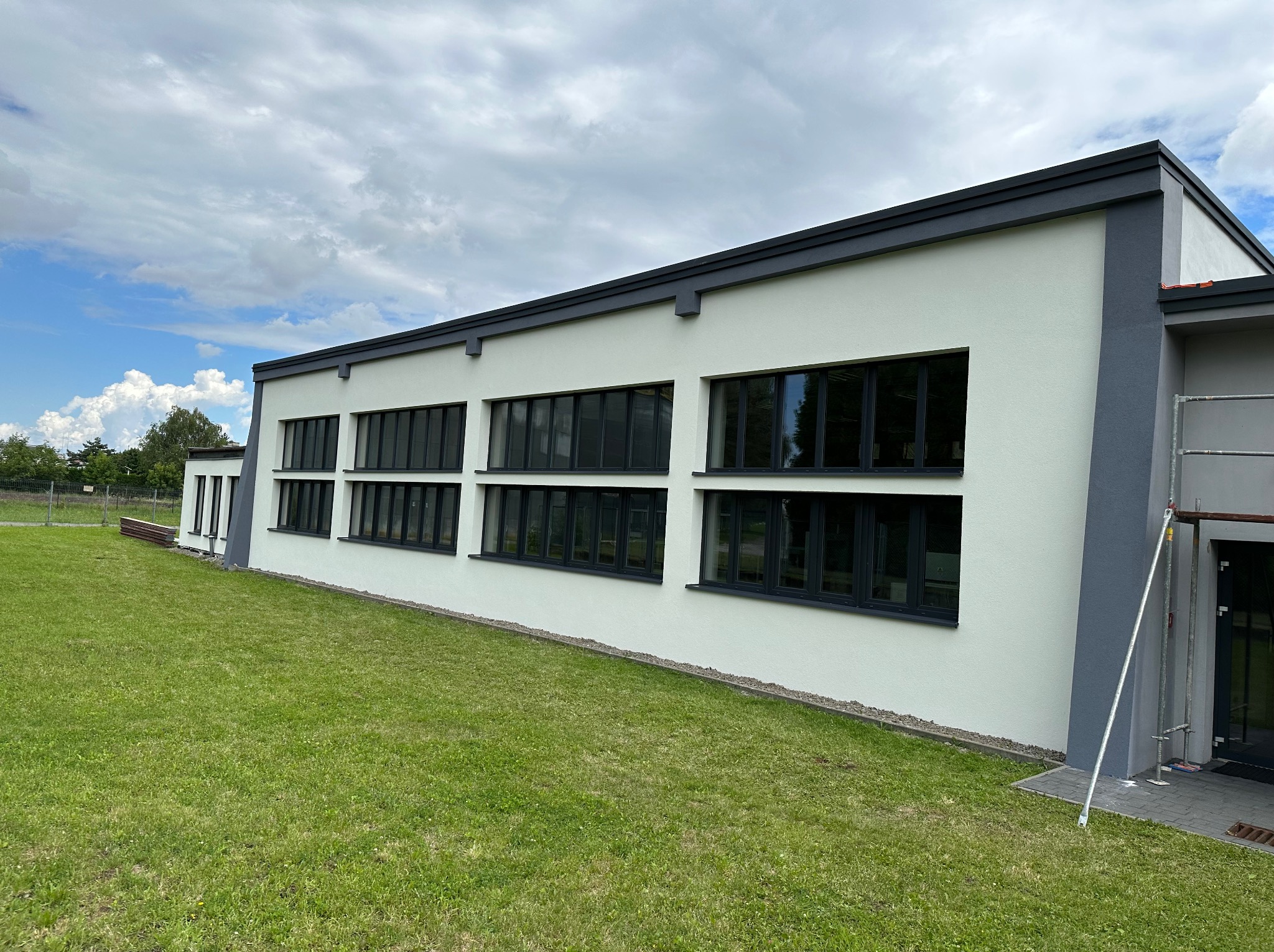 Świeżo otynkowany budynek z czarnymi ramami okiennymi i szarymi elementami wykończeniowymi, widok z poziomu trawnika, fragment rusztowania przy wejściu.