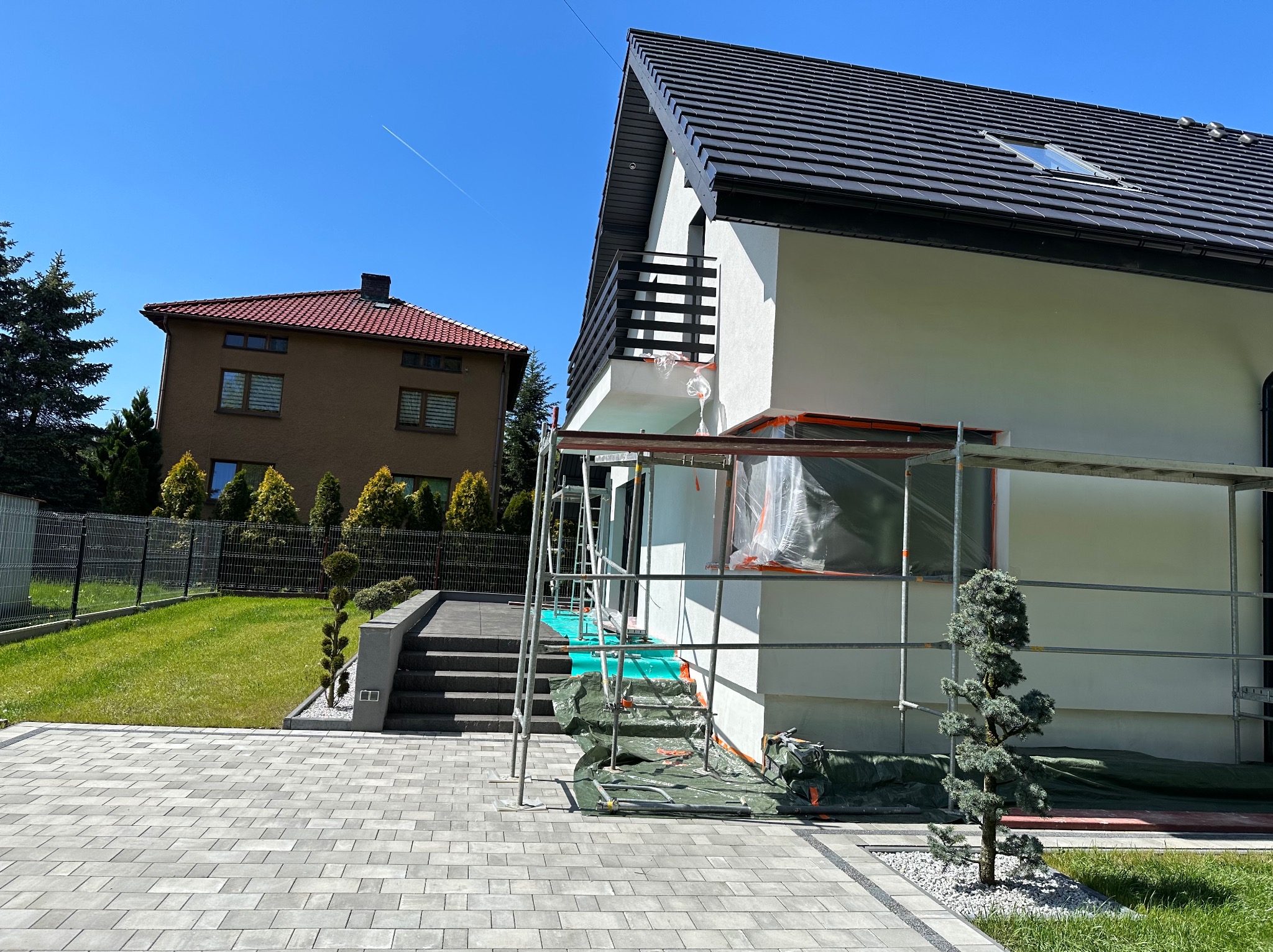 Dom jednorodzinny w trakcie prac elewacyjnych: biała fasada, czarna dachówka, rusztowania, zabezpieczone okna folią, widok na kostkę brukową i sąsiedni budynek pod błękitnym niebem.