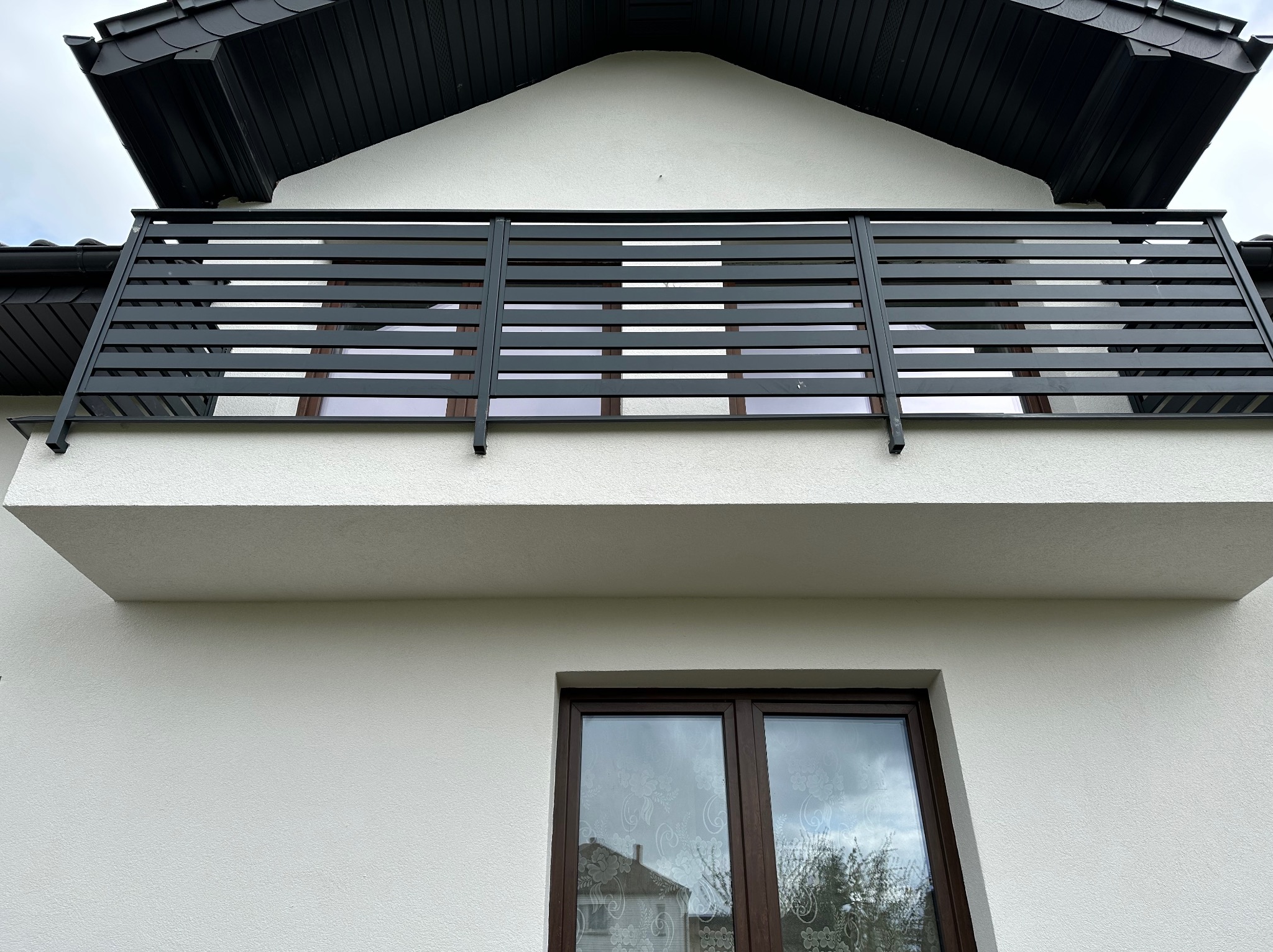 Biała elewacja domu z czarnym dachem i balkonem z poziomymi barierkami, okno z ciemną ramą odbija sąsiedni budynek.