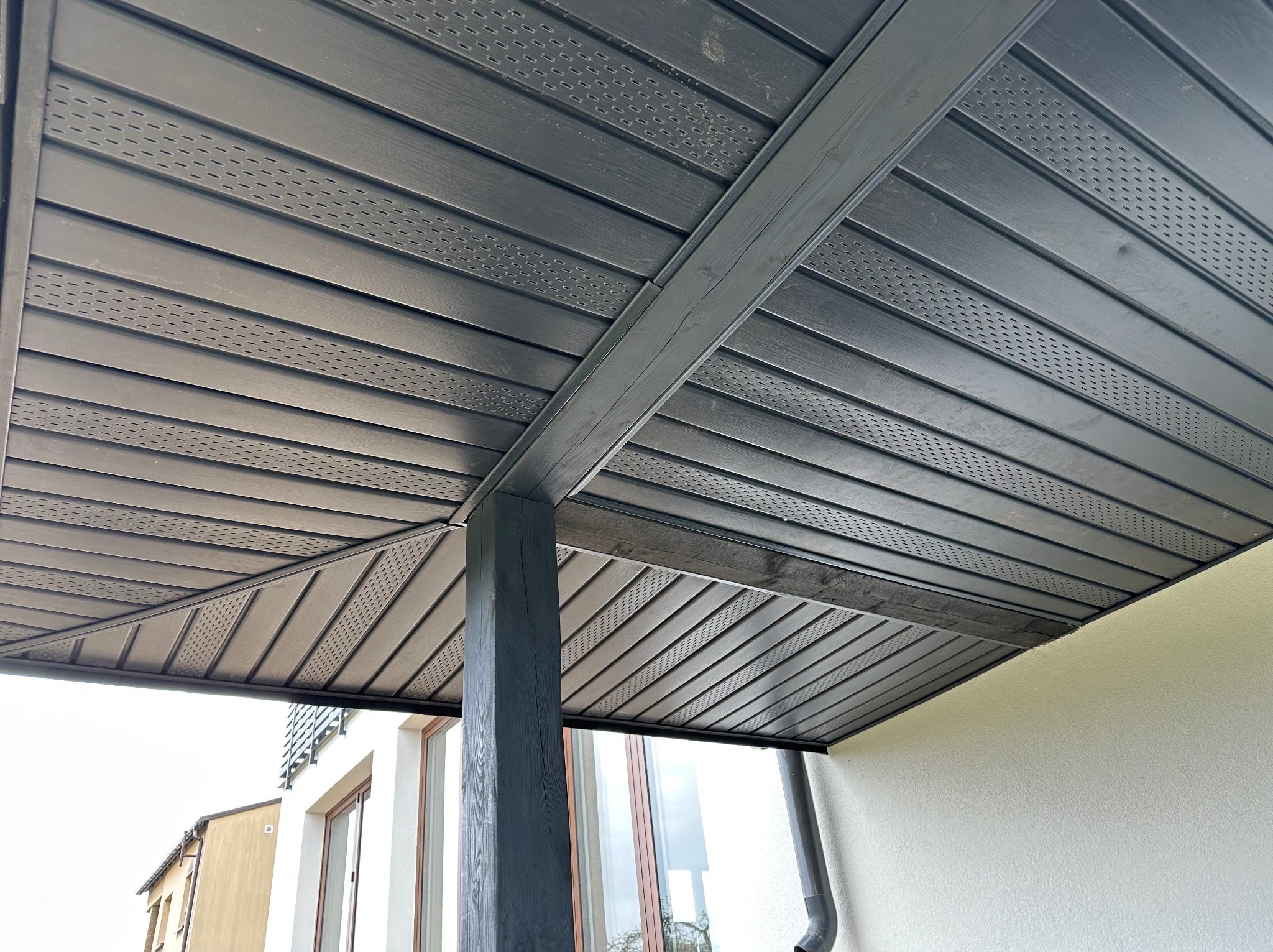 Nowoczesna, ciemnoszara podbitka dachowa z perforacją zamontowana na zewnątrz budynku, widok od dołu, słup podtrzymujący konstrukcję.