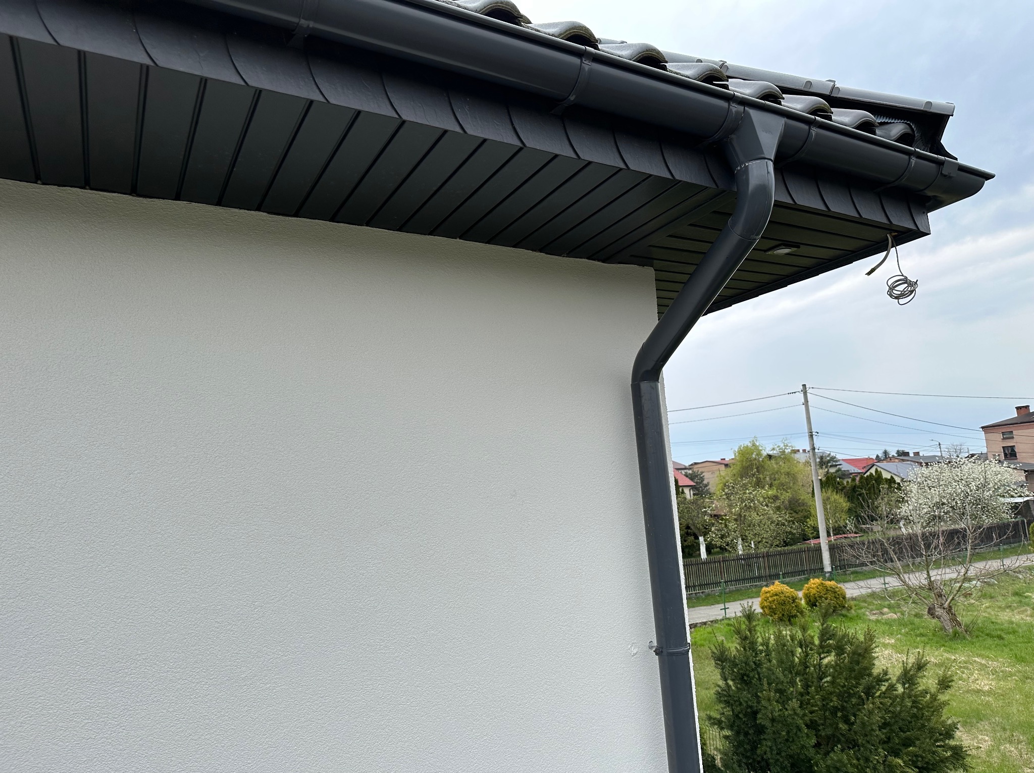 Fragment domu z jasną, chropowatą elewacją, ciemnoszarą podbitką dachową i rynną; widoczne zwisające kable elektryczne.