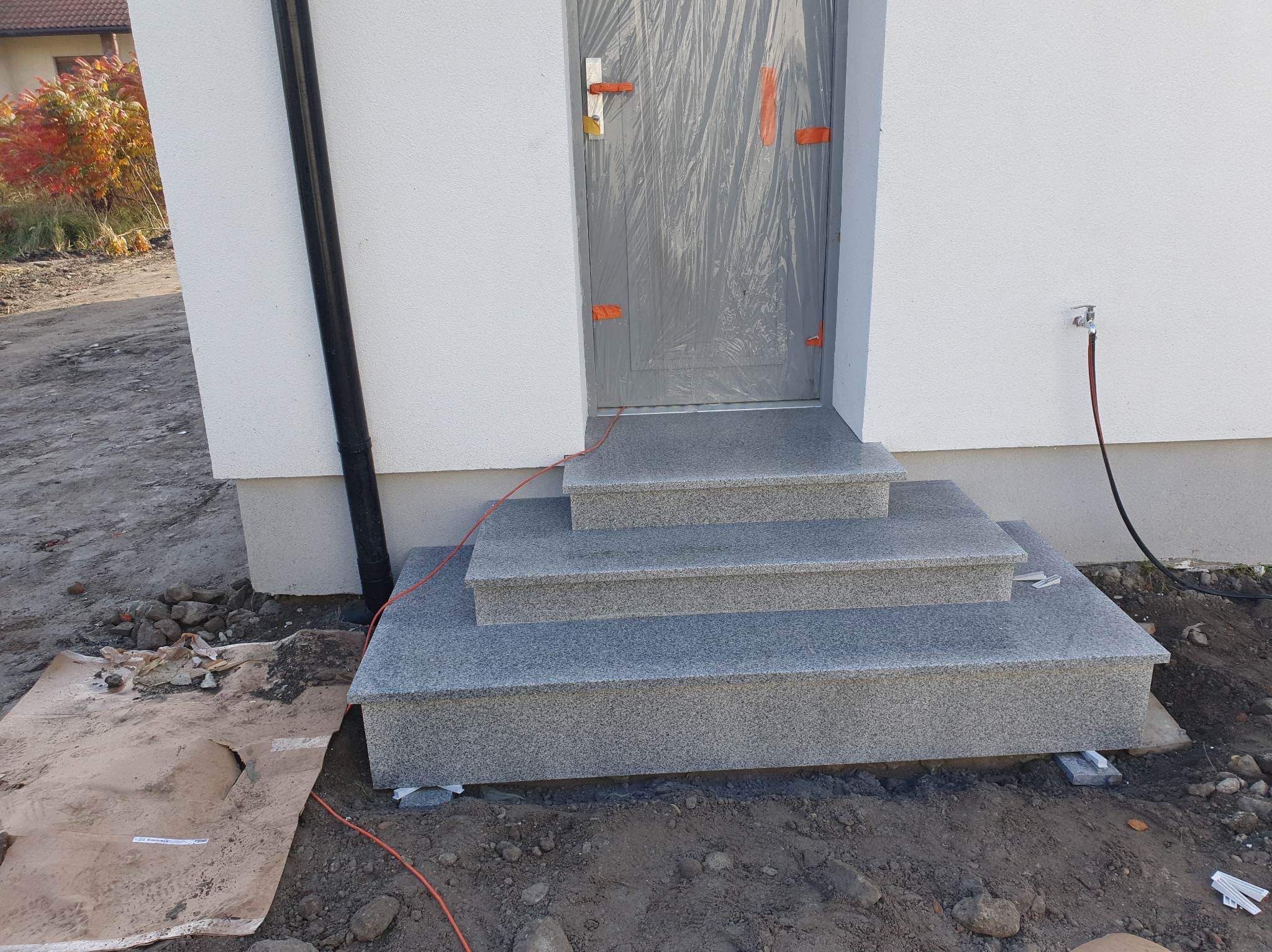 Nowe, granitowe schody wejściowe do domu, zabezpieczone folią drzwi i świeżo ułożone otoczenie z ziemią i gruzem.