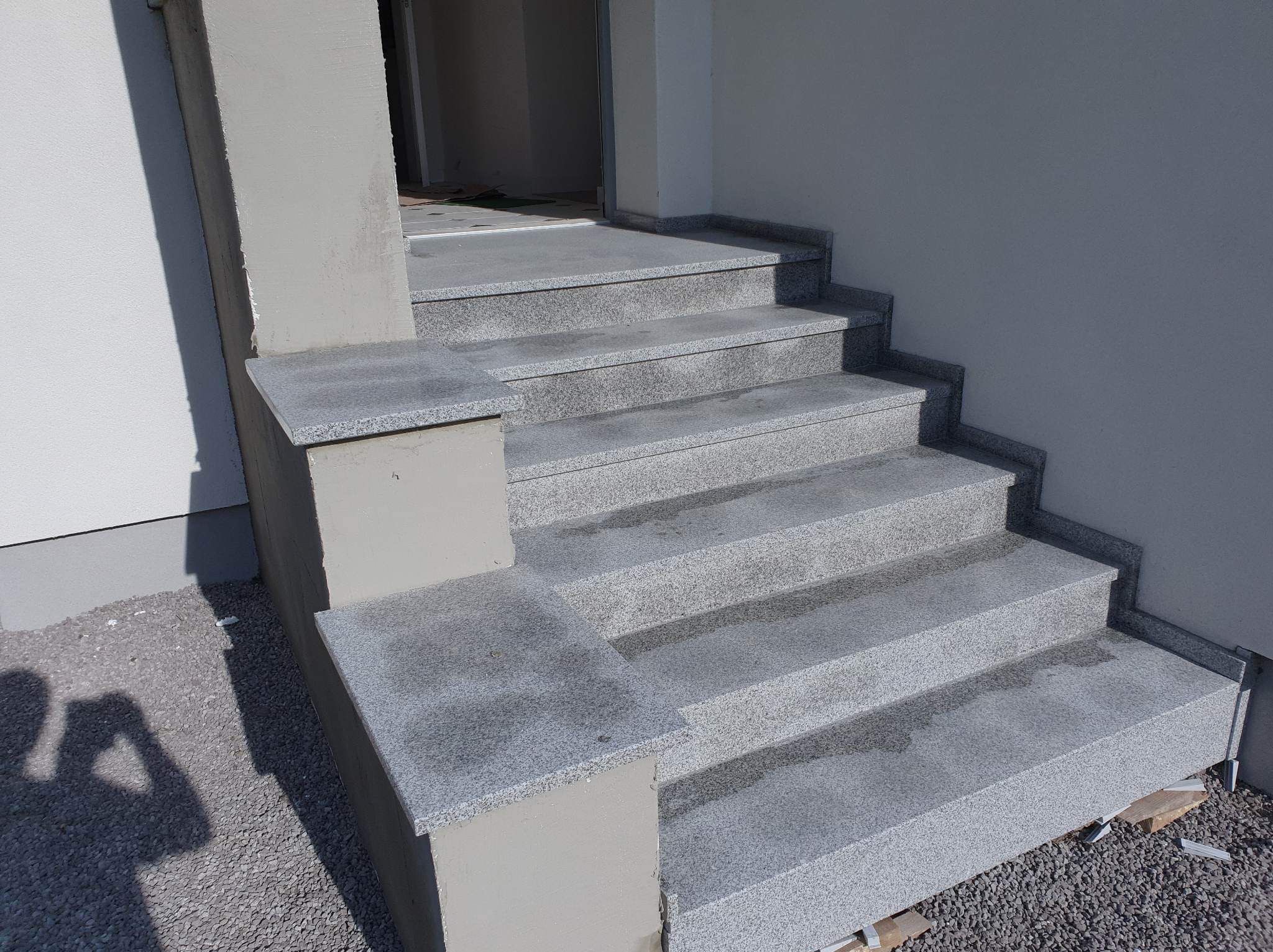 Granitowe schody wejściowe do budynku, z widocznymi elementami wykończeniowymi i szarym tynkiem na ścianie.