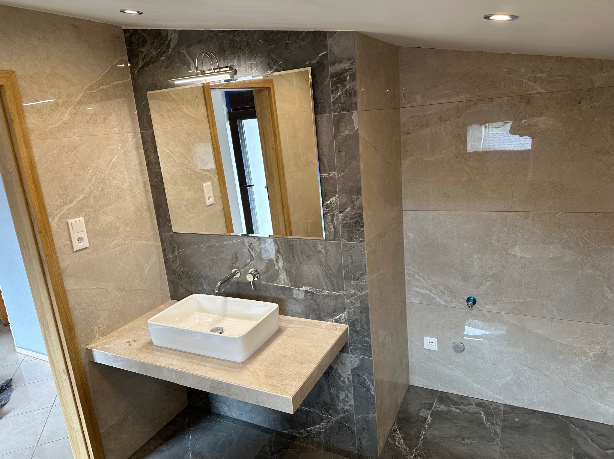 Nowoczesna łazienka z marmurowymi płytkami w odcieniach szarości i beżu, biała, prostokątna umywalka nablatowa i chromowana bateria, lustro z oświetleniem nad umywalką.