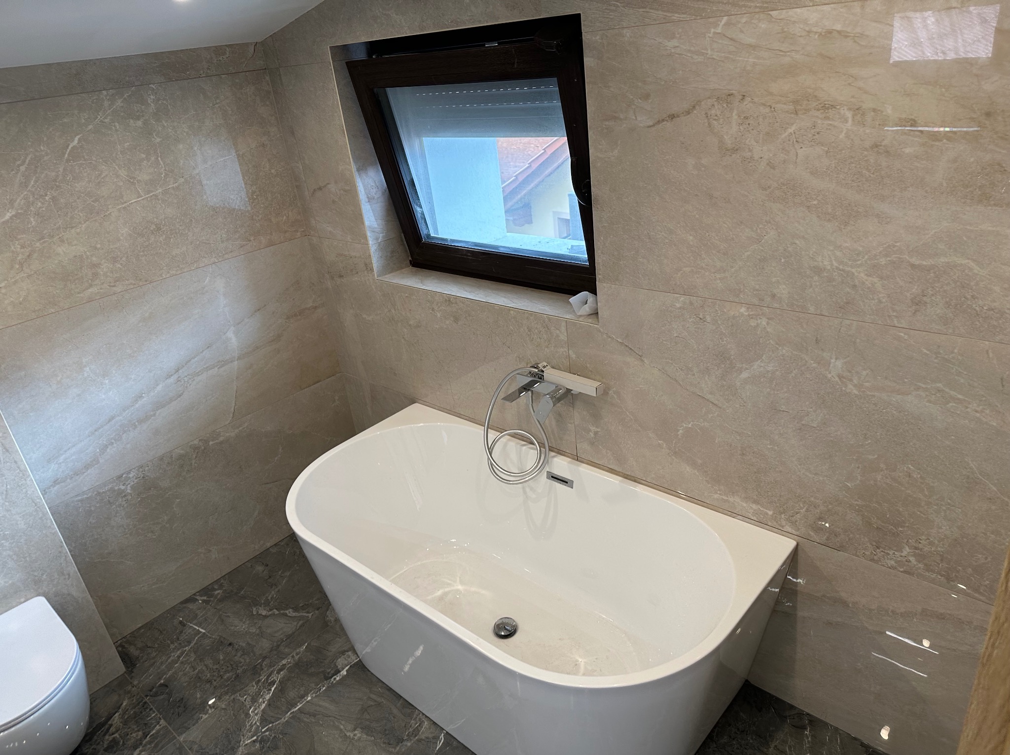 Nowoczesna łazienka na poddaszu z wolnostojącą białą wanną, szarymi marmurowymi płytkami podłogowymi i beżowymi płytkami ściennymi, okno dachowe z widokiem na dach sąsiedniego budynku.