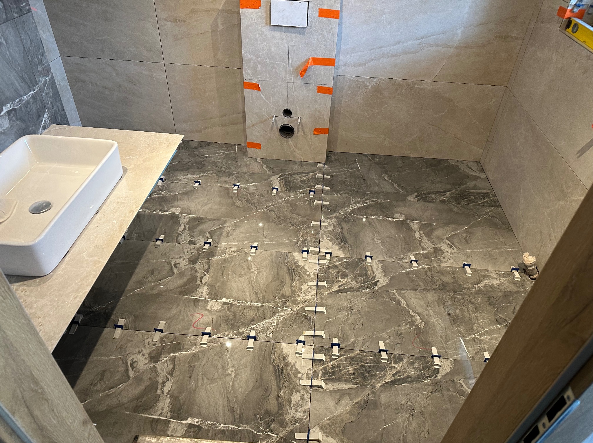 Montaż płytek podłogowych o marmurkowym wzorze w łazience, widoczne krzyżyki dystansowe, instalacja podtynkowa WC z pomarańczową taśmą zabezpieczającą.