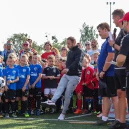 Trener demonstruje technikę kopnięcia piłki grupie dzieci z drużyny piłkarskiej w niebieskich koszulkach z logo sponsora, na tle zgromadzonego tłumu rodziców i innych osób.