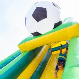 Chłopiec zjeżdża z dmuchanej zjeżdżalni w kształcie piłki nożnej, widok od dołu na jasnym tle nieba.