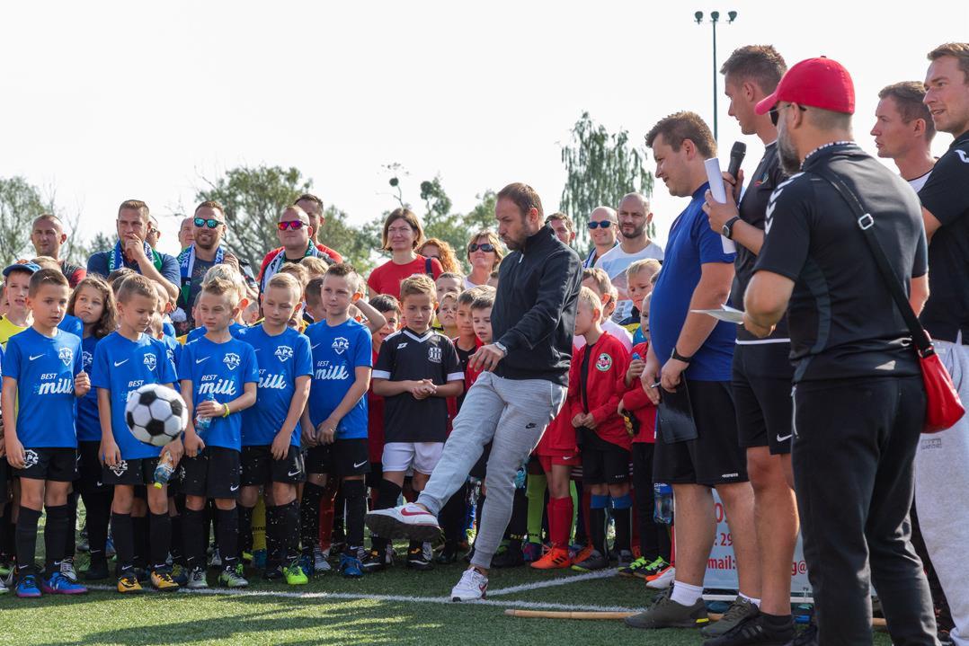 Trener demonstruje technikę kopnięcia piłki grupie dzieci z drużyny piłkarskiej w niebieskich koszulkach z logo sponsora, na tle zgromadzonego tłumu rodziców i innych osób.