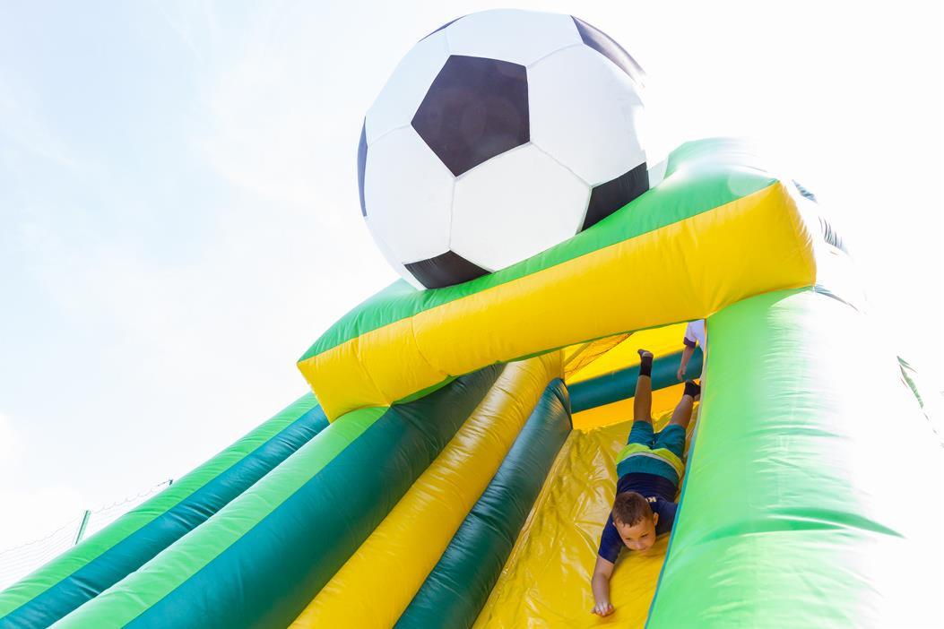 Chłopiec zjeżdża z dmuchanej zjeżdżalni w kształcie piłki nożnej, widok od dołu na jasnym tle nieba.