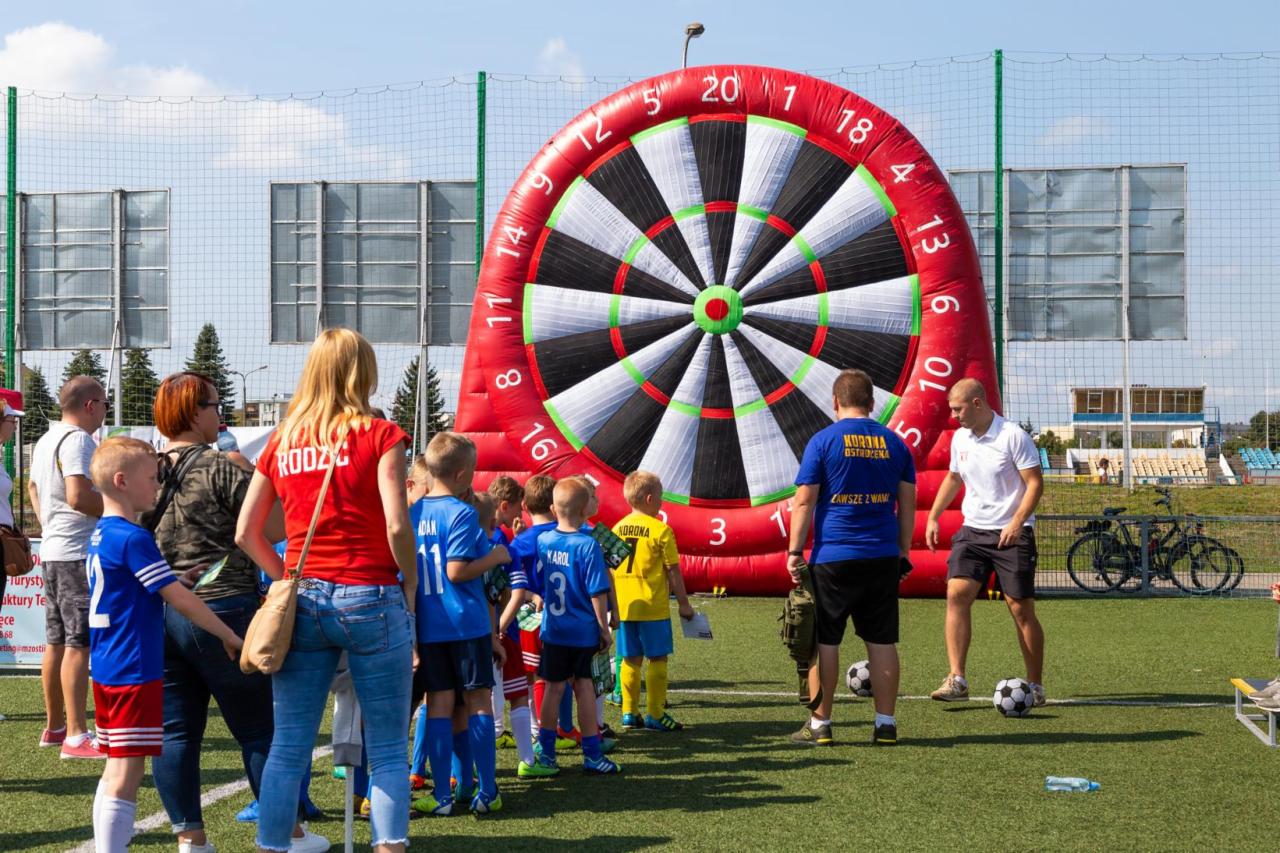 Grupa dzieci i dorosłych na trawiastym boisku, przed dmuchaną tarczą do darta w kolorach czerwonym, białym i czarnym, z widocznymi numerami, w tle siatka i budynek stadionu.