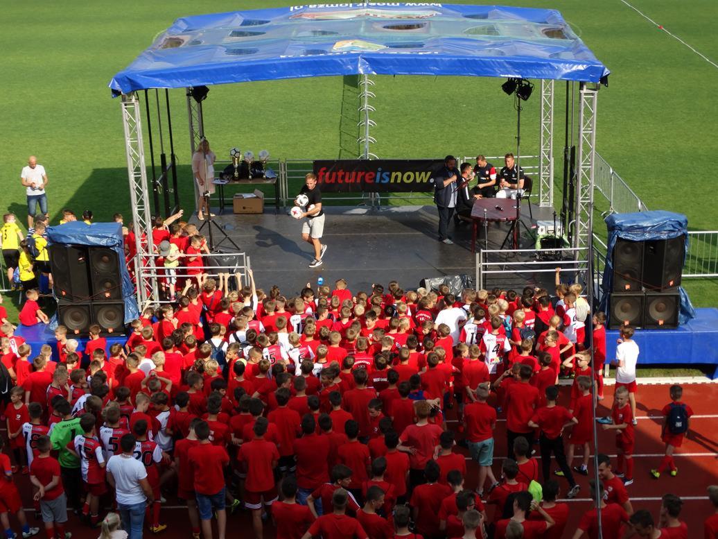 Duża grupa dzieci w czerwonych koszulkach zgromadzona przed sceną z nagłośnieniem i transparentem 'futureisnow.', na scenie stoi mężczyzna z piłkami do gry w rękach.