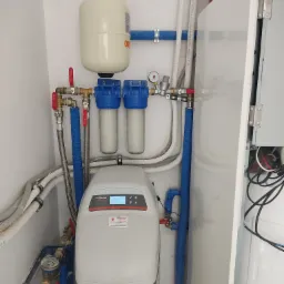 Zestaw uzdatniania wody Viessmann z filtrami mechanicznymi, zbiornikiem ciśnieniowym i rurami w niebieskiej izolacji, zamontowany przy białej ścianie i obudowie kotła.