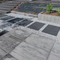 Nowoczesna aranżacja ścieżki w Jaworznie z dużych, szarych płyt betonowych i czarnych płyt oddzielonych białym grysem, prowadzącej do schodów i rabaty z roślinami.