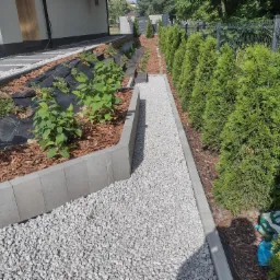 Ścieżka z białego grysu oddzielona szarą obrzeżem betonowym, prowadząca wzdłuż rzędu tuj, z widocznym fragmentem domu i ogrodu z korą oraz agrowłókniną.
