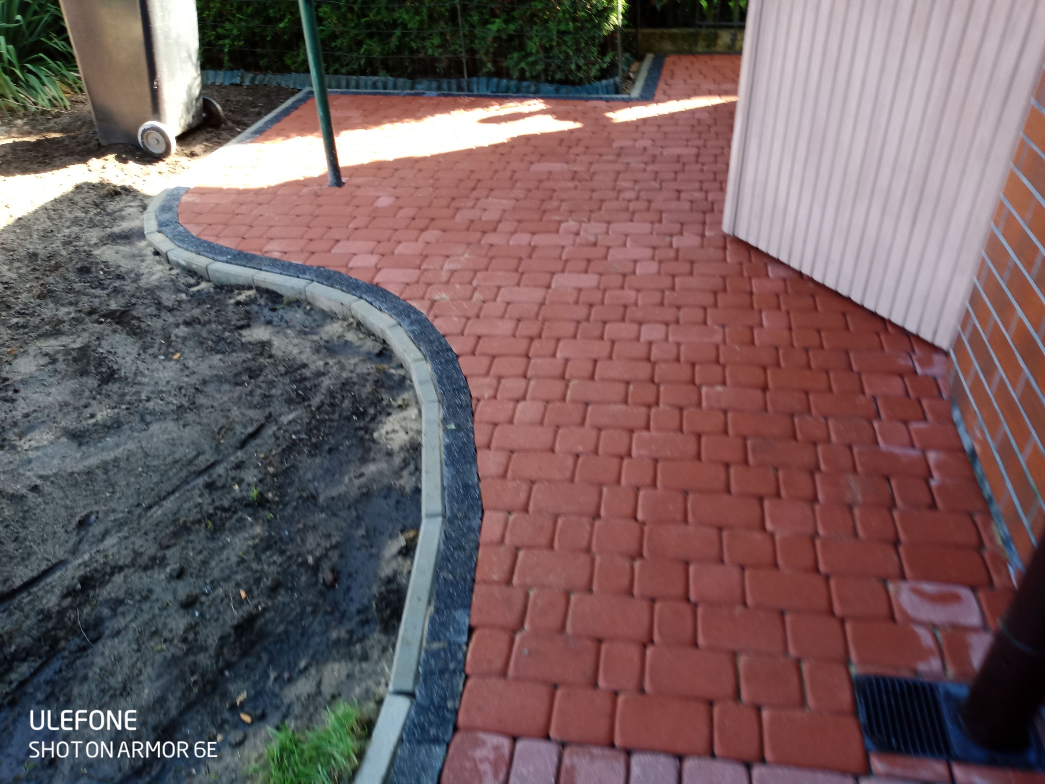 Czerwona kostka brukowa ułożona na posesji, widoczny fragment ogrodu z czarną ziemią i obrzeżem, fragment garażu i rynny.
