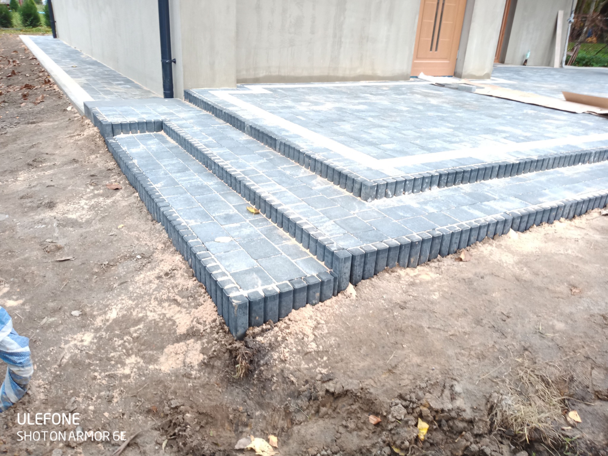 Nowo ułożone schody z ciemnej kostki brukowej prowadzące do domu, widoczne białe fugi i niedokończone otoczenie z ziemią.