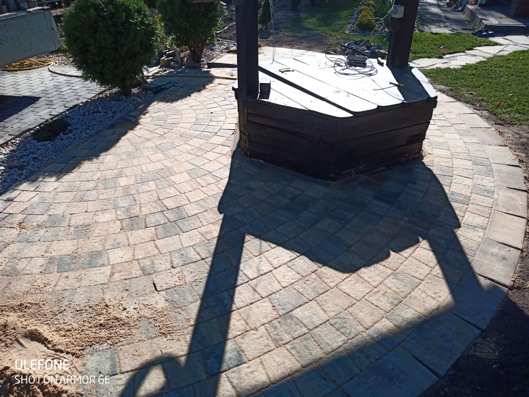 Studnia otoczona okrągłym wzorem z kostki brukowej w odcieniach szarości i beżu, zasypana piaskiem, z widocznym cieniem konstrukcji studni