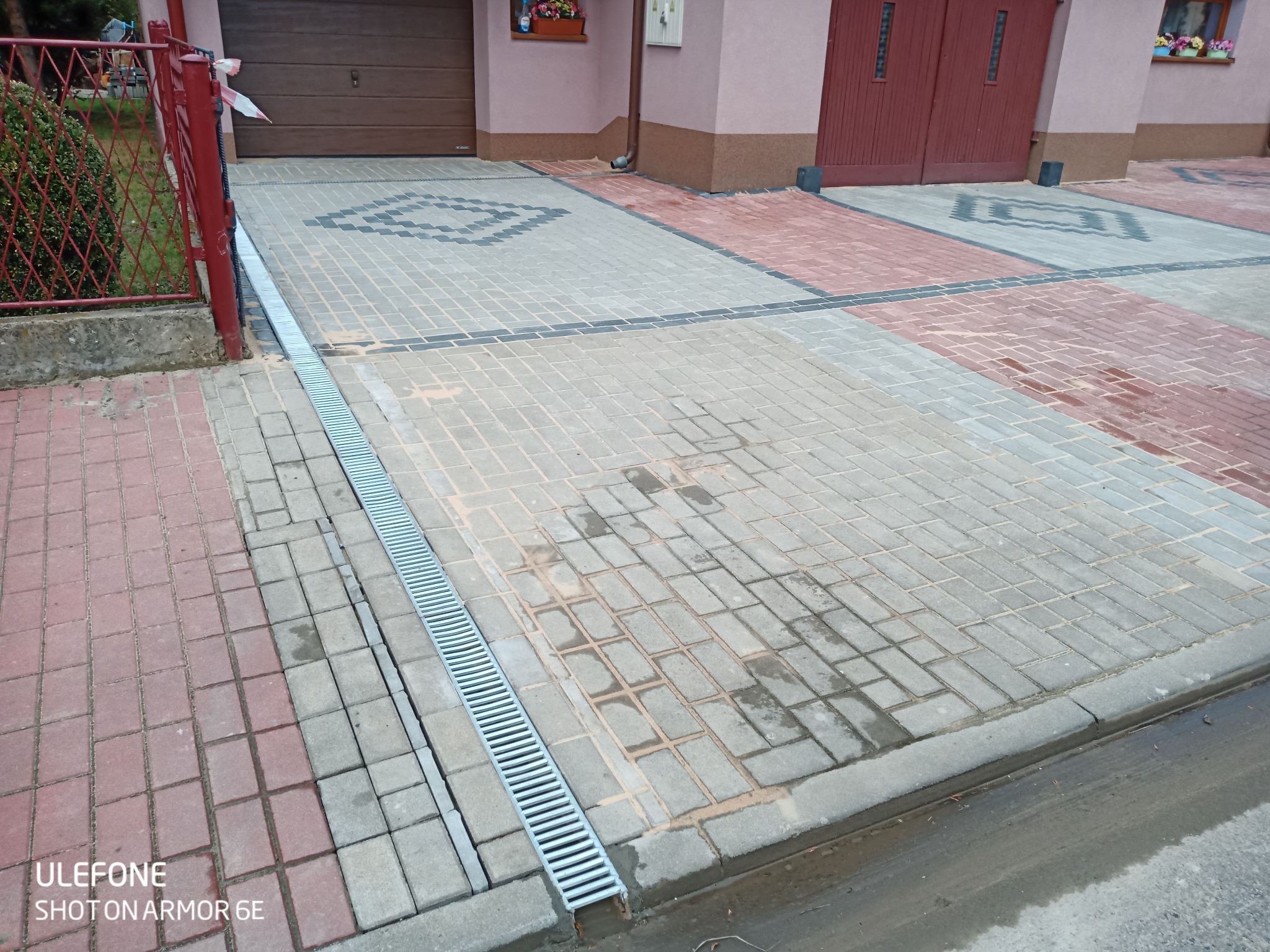 Ułożona kostka brukowa na podjeździe z odpływem liniowym, widoczne wzory z kostki w kolorach szarości i czerwieni, częściowo zabrudzona piaskiem.