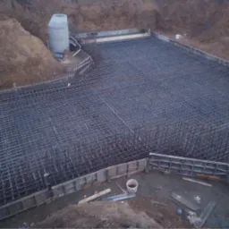 Siatka zbrojeniowa ułożona w wykopie, przygotowana do zalania betonem, z widocznymi szalunkami i elementami konstrukcyjnymi w tle.