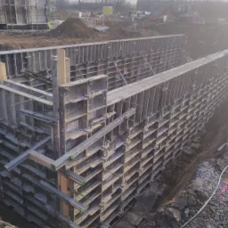Szare szalunki stalowe tworzące formę dla fundamentów, widoczne zbrojenia i wykop, w tle budynek w trakcie budowy i koparka.