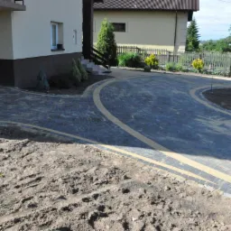 Ułożona ciemnoszara kostka brukowa z jasnymi pasami tworząca ścieżkę i podjazd przed domem z elementami zieleni i świeżo nasadzonej roślinności.