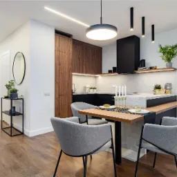 Nowoczesna kuchnia z drewnianymi frontami, czarnymi akcentami i wyspą połączoną z drewnianym stołem jadalnianym oraz szarymi krzesłami.