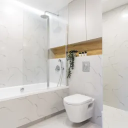 Nowoczesna łazienka z białymi marmurkowymi płytkami, wiszącą toaletą, szklanym parawanem nawannowym i drewnianą wnęką z roślinami.