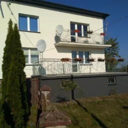 Dwukondygnacyjny dom z jasną elewacją, balkonami z kwiatami i dwiema antenami satelitarnymi, widoczny z perspektywy ogrodu z tujami i kamiennym ogrodzeniem.