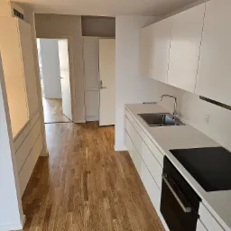 Nowoczesna, minimalistyczna kuchnia z białymi szafkami i drewnianą podłogą w Nowym Sączu, widok na otwarte drzwi do innego pomieszczenia.