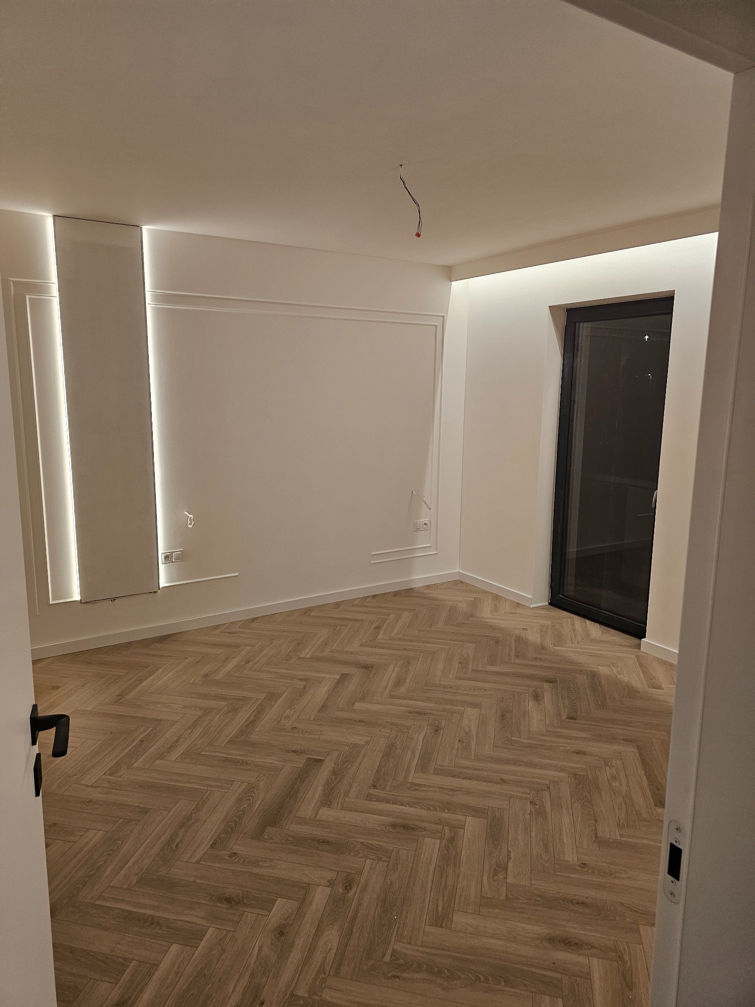 Jasne wnętrze z parkietem w jodełkę, listwami dekoracyjnymi i oświetleniem LED. Widoczne gniazdka elektryczne i wystające kable. Minimalistyczny design.