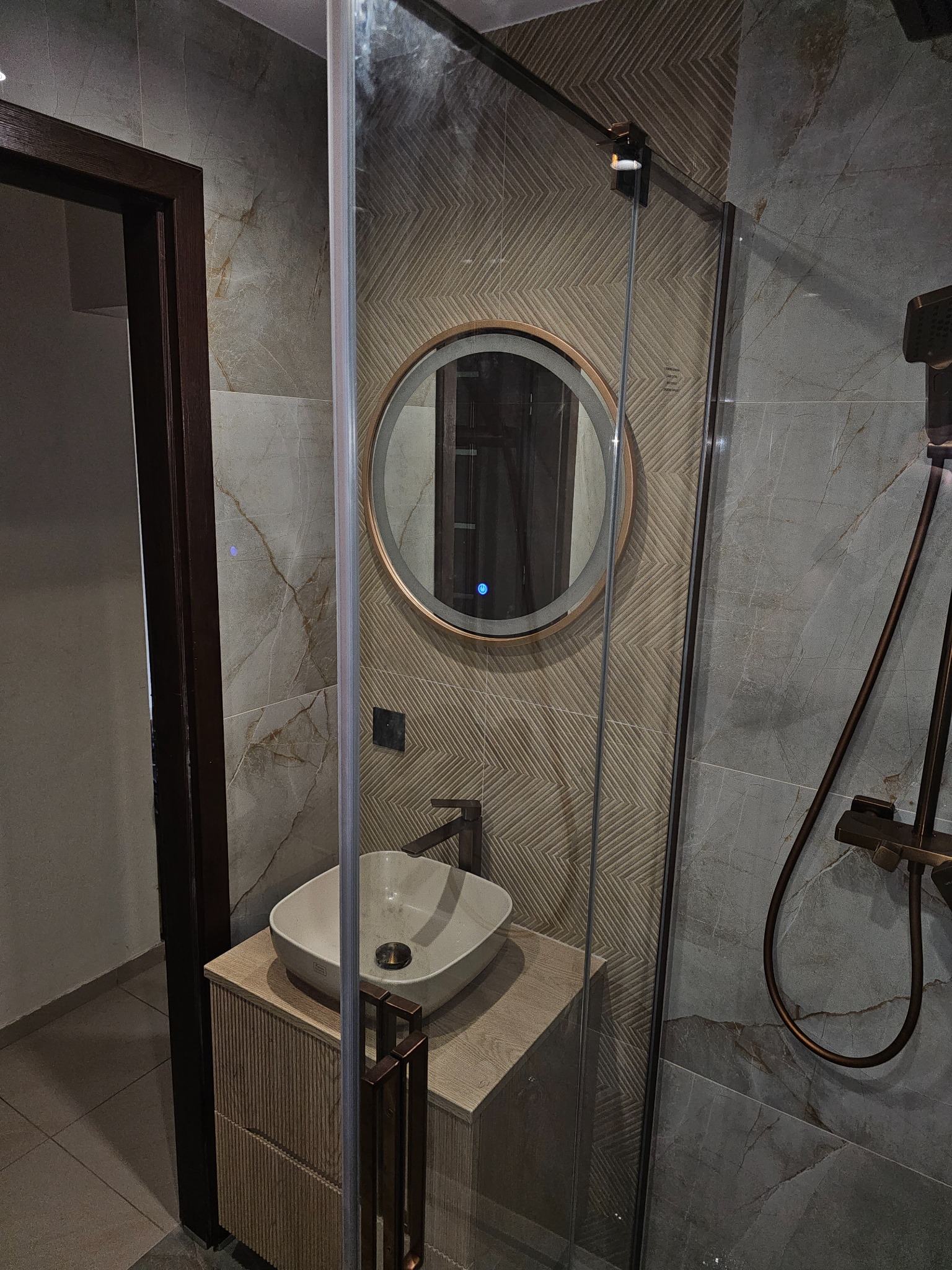 Nowoczesna łazienka z okrągłym lustrem LED, umywalką nablatową i kabiną prysznicową. Widoczne płytki imitujące marmur i geometryczny wzór na ścianie.