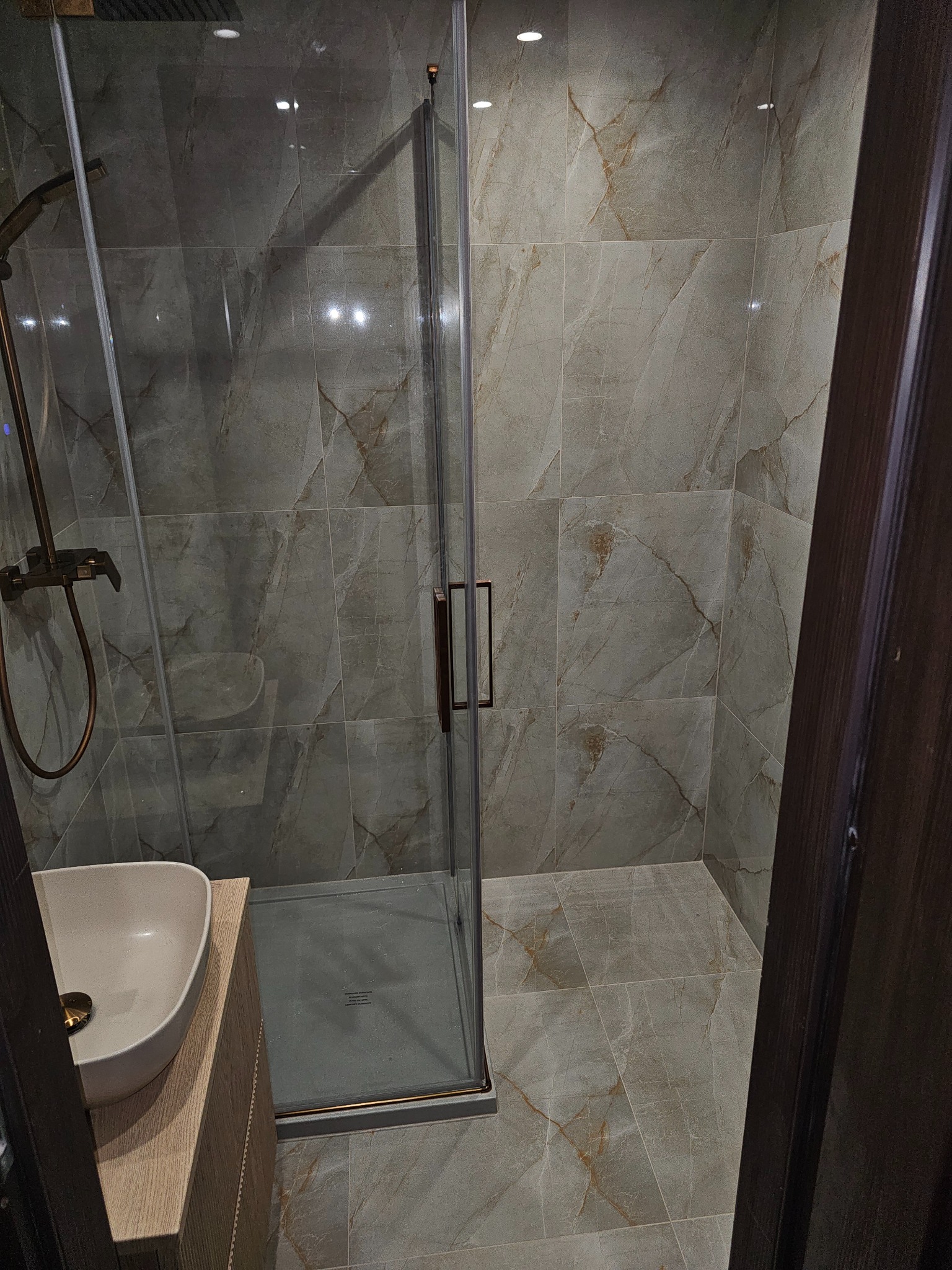 Wykończona łazienka z kabiną prysznicową, umywalką nablatową i płytkami imitującymi marmur w odcieniach szarości i beżu. Armatura w kolorze starego złota.