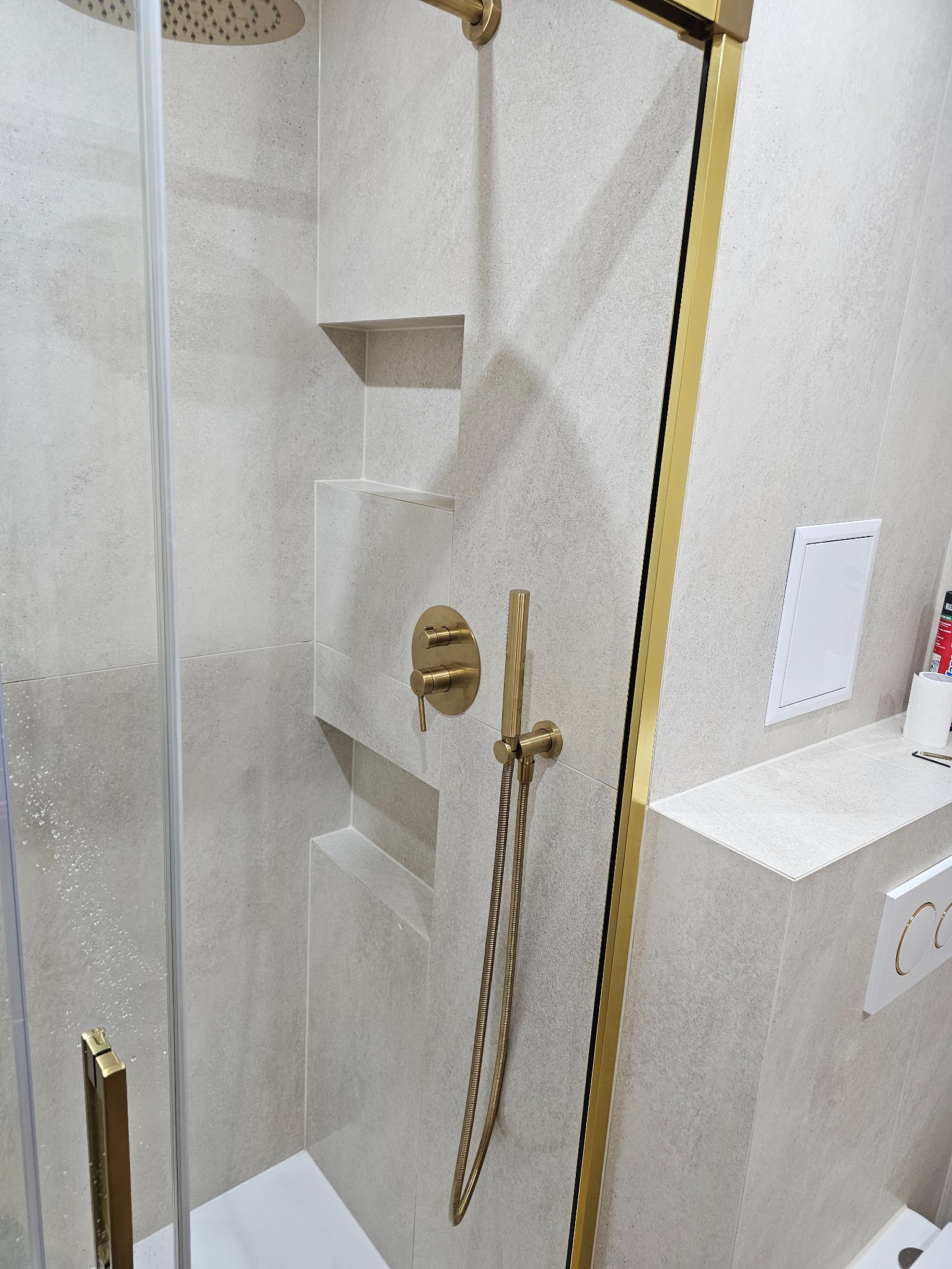 Beżowa kabina prysznicowa z złotymi detalami: rama, bateria, słuchawka. W tle widoczne półki w ścianie i przyciski spłuczki. Minimalistyczny design.