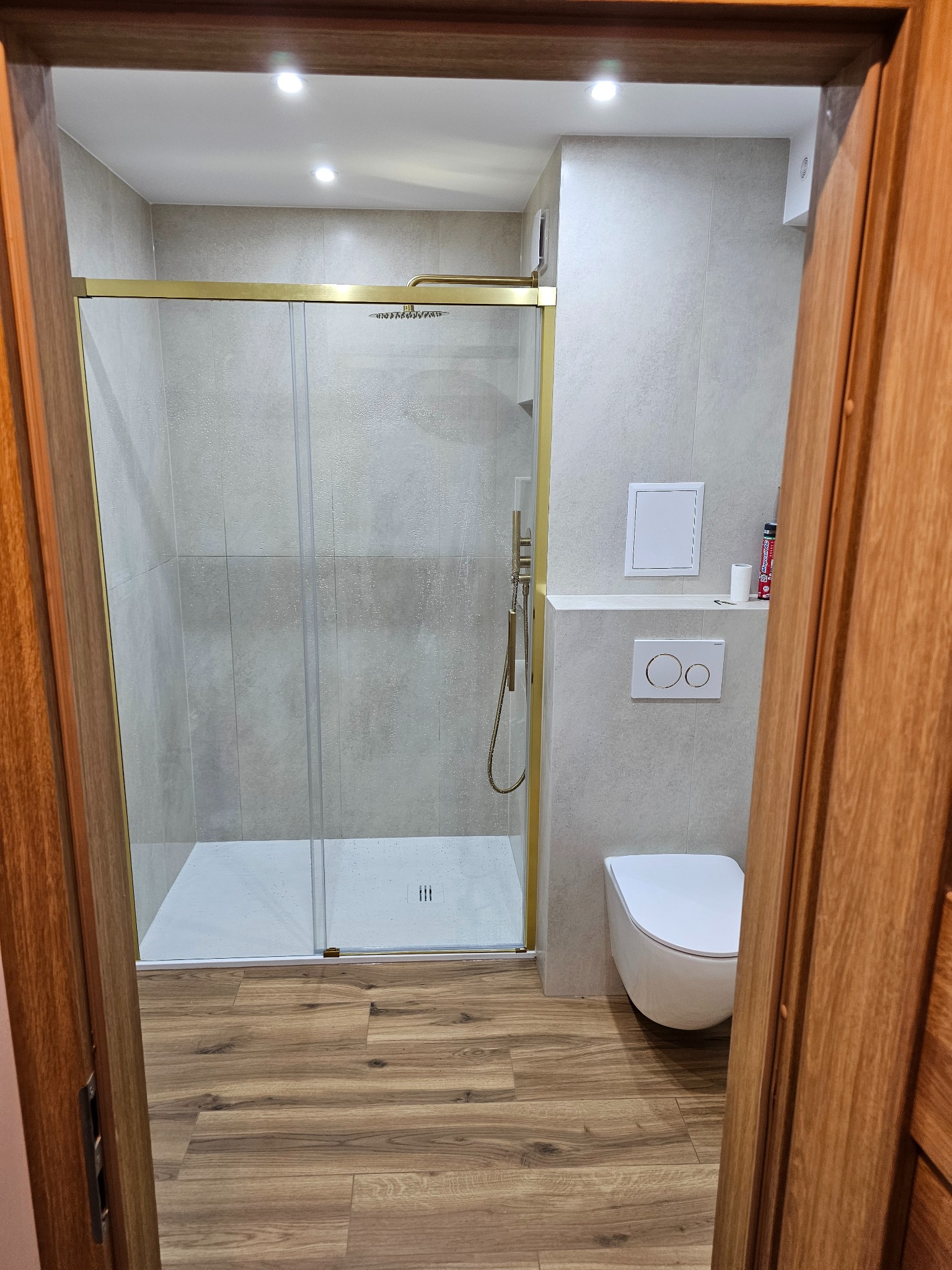 Wykończona łazienka z kabiną prysznicową w złotej ramie, podwieszaną toaletą i panelami podłogowymi imitującymi drewno, widziana zza drewnianej ościeżnicy.