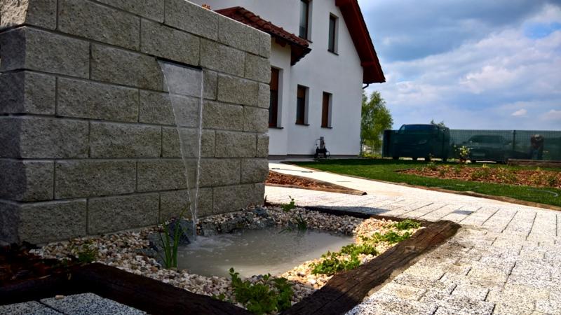 Ogród z kaskadą wodną spływającą po murze z szarej kostki, mały staw z kamieniami i roślinami, ścieżka z jasnej kostki brukowej, dom z białymi ścianami w tle.